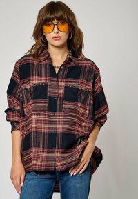 Chemise en flanelle à carreaux noirs et rouges avec fermeture par boutons, deux poches poitrine avec clous, coupe décontractée, manches longues, portée sur un jean.