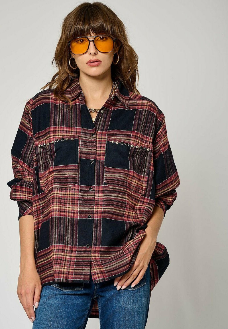 Chemise en flanelle à carreaux noirs et rouges avec fermeture par boutons, deux poches poitrine avec clous, coupe décontractée, manches longues, portée sur un jean.