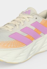 Close-up van een lichtbeige sneaker met roze strepen en oranje accenten, met crèmekleurige veters en een dikke witte zool.