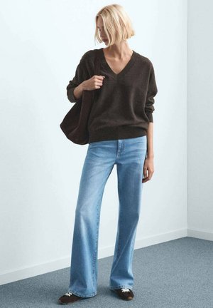 Pulover maro închis cu guler în V, croială lejeră, purtat cu blugi evazați bleu deschis. Ținuta este accesorizată cu o geantă tote închisă la culoare și pantofi maro.