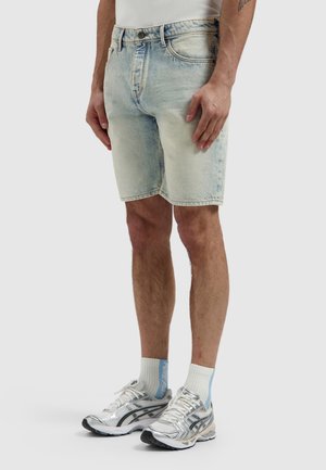 Man draagt lichtblauwe denim shorts, witte sokken met blauwe tekst en zilverkleurige sportschoenen, staand tegen een effen achtergrond.