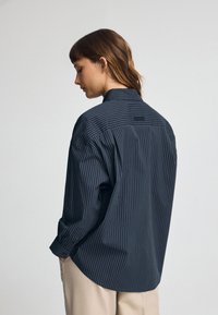 Granatowa, paskowana koszula zapinana na guziki o luźnym kroju, z długimi rękawami i detalem z tyłu. Materiał wydaje się gładki o lekkiej teksturze.