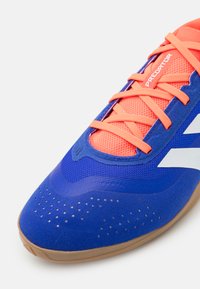Zapatilla deportiva azul con parte superior texturizada, cordones naranjas y acentos blancos. Presenta una suela de goma y detalles perforados cerca de la puntera.