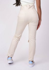 Beige high-waisted broek met een aansluitend ontwerp, twee achterzakken en een gladde textuur. Gekenmerkt door lichte sneakers.