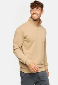 Pull beige avec un col montant et une fermeture éclair. Tissu doux et texturé avec des poignets et un ourlet côtelés. Présente un logo brodé sur la poitrine.