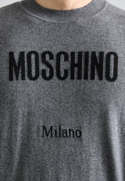 Pilkas megztinis su „MOSCHINO“ užrašu didelėmis juodomis raidėmis ir „Milano“ mažesniu šriftu. Minkšta tekstūra ir apvalio kaklo dizainas.