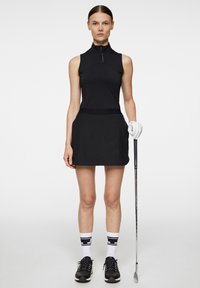 Svart ärmlös golftopp med hög krage, kombinerad med en svart skort. Modellen håller en golfklubba, bär svarta skor och vita strumpor med svarta ränder.