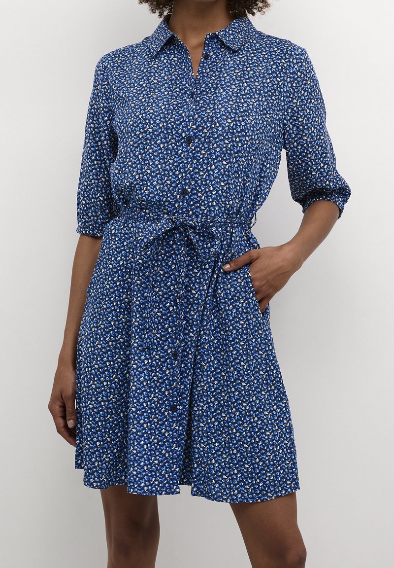 Femme portant une robe courte bleue avec un petit imprimé floral, boutonnée devant, manches trois-quarts et ceinture nouée à la taille.