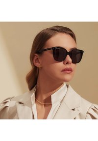 Schwarze rechteckige Sonnenbrillen mit dunklen Gläsern haben eine glänzende Oberfläche und goldene Logoakzente. Das Modell trägt einen hellen Blazer und eine Halskette.