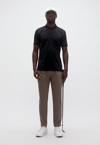 Άνδρας που φοράει μαύρο πόλο, μπεζ παντελόνι, άσπρο καπέλο και παπούτσια, κρατώντας ένα μπαστούνι γκολφ μπροστά σε ένα απλό, φωτεινό φόντο.