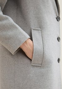 Manteau en laine gris avec une texture lisse, doté d'une fermeture à un seul bouton, d'une poche latérale et d'une coupe ajustée.