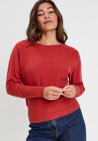Jersey de punto rojo con cuello redondo y puños y dobladillo acanalados, que presenta una textura suave y un diseño ajustado, combinado con unos jeans azul oscuro.