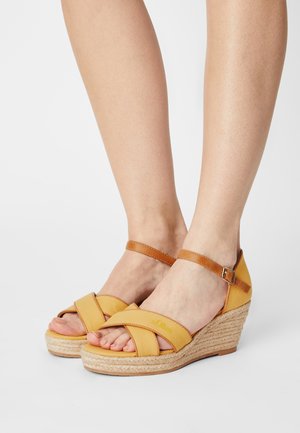 Yellow wedge sandals with crossed fabric straps, leather ankle strap, and woven jute platform. Features a low heel and open toe design.