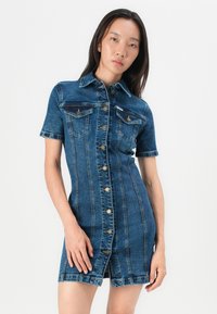 Robe en denim avec des manches courtes, une fermeture à boutons sur le devant et deux poches poitrine. Bleu foncé avec des coutures visibles et une silhouette ajustée.