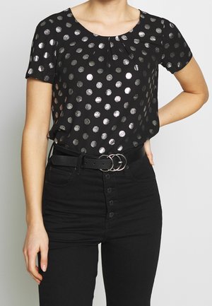 Femme portant un haut noir à manches courtes avec des pois argentés, rentré dans un pantalon noir taille haute avec une ceinture noire avec deux anneaux argentés.