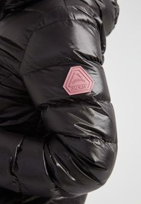 Primer plano de la manga de una chaqueta acolchada negra y brillante con un parche hexagonal elevado de color rosa que dice "JACK1T" en la parte superior del brazo.