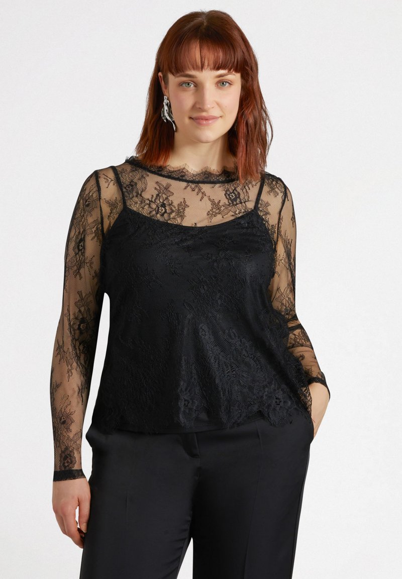 Top in pizzo nero con maniche lunghe trasparenti, sovrapposto a una canottiera nera a tinta unita. Presenta motivi floreali e bordi a scallop lungo il décolleté.