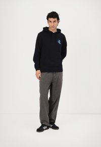 Juodas hoodie su mėlynu grafiniu dizainu ant krūtinės, pilki platūs keliai ir juodi batai su baltais dryžiais. Paprastas, kasdienis įvaizdis.
