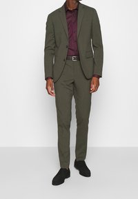 Olivově zelený oblek na míru se strukturovaným sakem a slim-fit kalhotami. Doplňený burgundskou košilí na knoflíky a černými botami.
