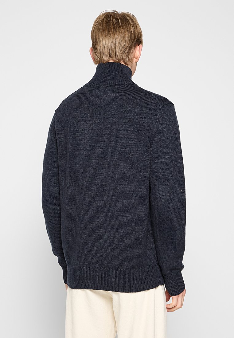 Mężczyzna w granatowym swetrze z golfem i białych spodniach, widziany z tyłu na jasnym tle.