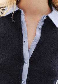 Cardigan bleu marine foncé avec une chemise à col bleu clair bouton-Down. Présente des détails de boutons et un tissu doux et texturé.