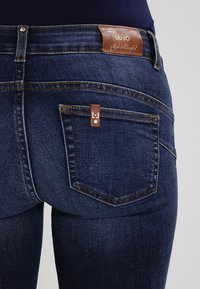 Jeans de mezclilla oscura con una etiqueta de cuero marrón en la cintura, un bolsillo trasero plano con una pequeña etiqueta marrón y detalles de costura sutiles.