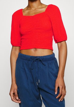 Femme portant un haut court texturé rouge vif avec des manches bouffantes jusqu'aux coudes et un pantalon bleu marine taille haute à cordon de serrage.