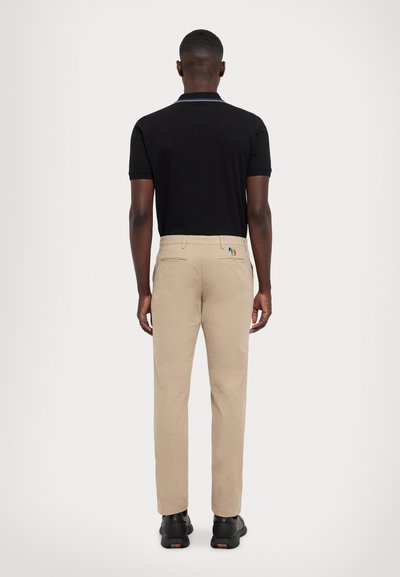 Calças bege com corte ajustado, bolsos traseiros e detalhes de costura em teal, amarelo e vermelho contrastantes. Combinadas com uma camisa polo preta.