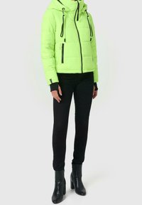 Giacca puffer verde neon con cappuccio, con dettagli neri, zip frontale e tasche laterali. Abbinata a pantaloni neri slim-fit e stivaletti.