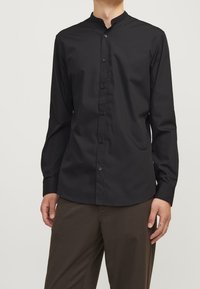 Svart långärmad skjorta med mandarin-krage, tillverkad av mjukt tyg, med knappar fram och en skräddarsydd passform. Buren med bruna byxor.