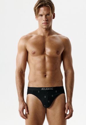 Umięśniony mężczyzna o blond włosach stoi przodem, ubrany w czarne majtki marki ATLANTIC na jednolitym białym tle.