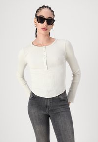 Svijetlo bež longsleeve rebrasti top s okruglim izrezom i zatvaranjem na pet dugmadi, uparen s tamnosivim uskim trapericama.