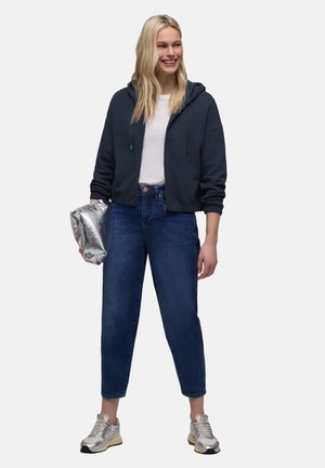 Femme souriante portant un sweat à capuche bleu marine, une chemise blanche, un jean bleu, des baskets argentées, tenant une pochette argentée, posant devant un fond blanc.