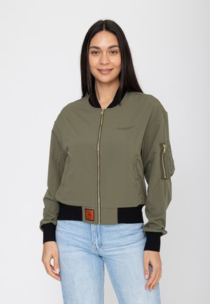 Kobieta z długimi ciemnymi włosami, ubrana w oliwkowo-zieloną kurtkę typu bomber z czarnymi mankietami oraz jasnoniebieskie dżinsy, uśmiecha się i stoi na białym tle.