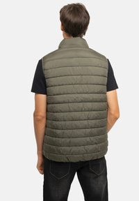 Veste sans manches matelassée olive verte avec un design matelassé horizontal, col montant, et matériau léger. Portée sur un t-shirt noir, vue de dos.