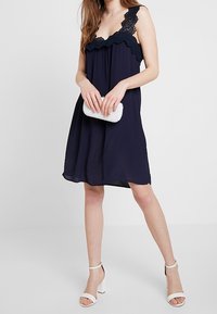 Kvinna klädd i en ärmlös marinblå klänning med spetsdetaljer, som håller en vit pärlclutch och bär vita klacksandaler framför en enfärgad bakgrund.