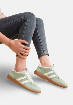 Personne portant des baskets vertes et blanches à lacets avec des semelles en caoutchouc, assise les jambes croisées, les mains reposant autour d'une cheville, vêtue d'un jean skinny foncé.