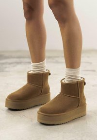 Botas de tobillo de ante marrón con una superficie texturizada, que cuentan con una gruesa suela plataforma, un forro suave y una lengüeta en la parte trasera para facilitar el uso.