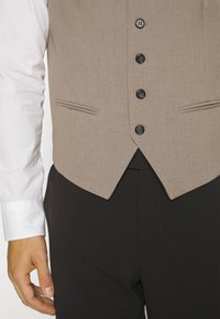 Beige väst med fem svarta knappar, två framfickor och en figursydd design. Bärs över en vit skjorta och matchas med svarta byxor.
