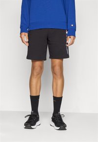 Pantaloni sportivi neri con una superficie liscia e un logo sul lato, abbinati a calze nere e scarpe da ginnastica nere. Maglia a maniche lunghe blu.