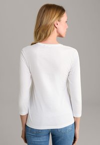 White long-sleeve shirt with a round neckline, soft texture, and subtle branding on the back. Paired with blue denim jeans.