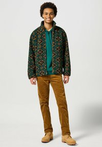 Chaquetón verde con un patrón de diamantes sobre una sudadera azul verdosa, combinado con pantalones de pana marrones y botas beige de cordones.