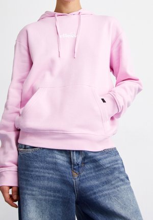 Osoba ubrana w jasnoróżową bluzę z kapturem z przednią kieszenią i sznurkami, w połączeniu z niebieskimi dżinsami, na jednolitym białym tle.