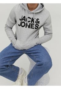 Šedá mikina s kapucí s černým logem "JACK & JONES", obsahuje klokaní kapsu a kapuci se stahovací šňůrkou, doplněná modrými džínami a bílými teniskami.