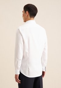 Camicia bianca a maniche lunghe con una vestibilità adattata, orlo arrotondato e polsini con bottoni, realizzata in tessuto di cotone morbido. Dettaglio di cucitura sottile sui polsini.