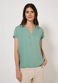 Blouse vert clair à manches courtes avec un décolleté en V et une texture douce et lisse. Coupe légèrement ample, froncée à l'ourlet pour un look décontracté.