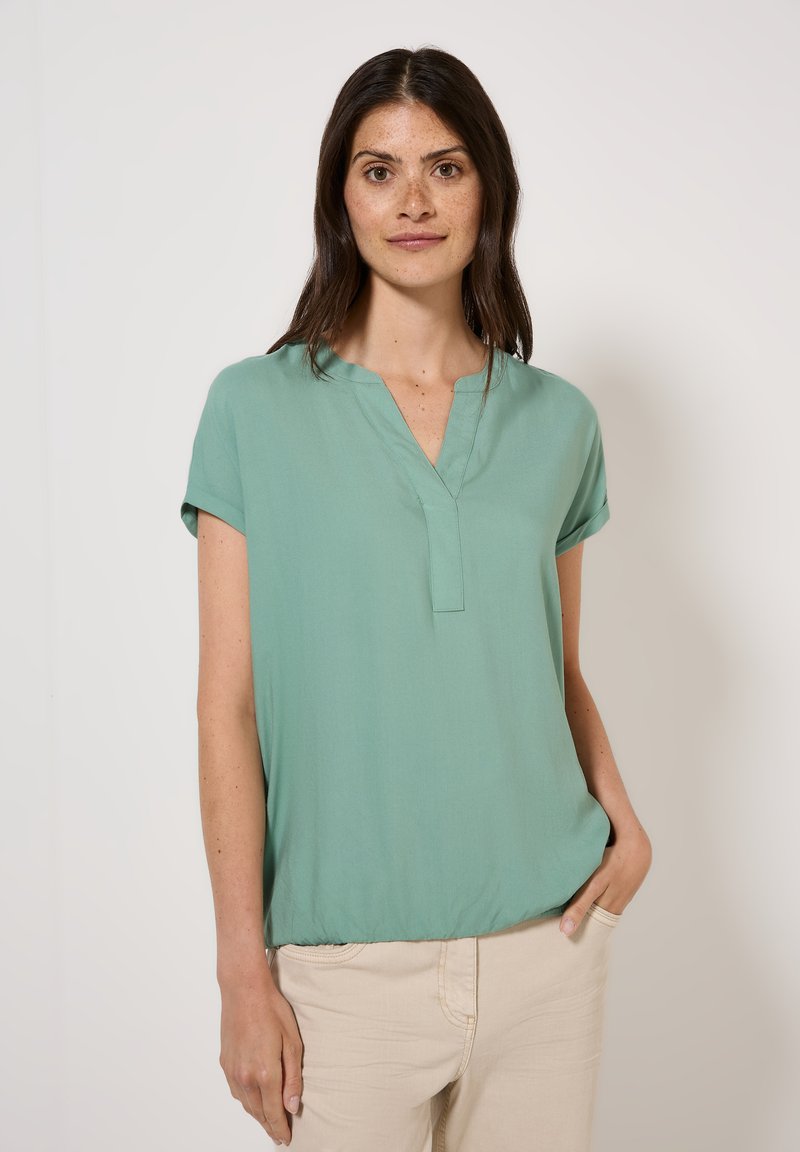 Blouse vert clair à manches courtes avec un décolleté en V et une texture douce et lisse. Coupe légèrement ample, froncée à l'ourlet pour un look décontracté.