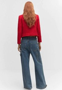 Cardigan court rouge en tissu texturé associé à un pantalon cargo en denim bleu à coupe décontractée, avec de grandes poches et une silhouette ample.