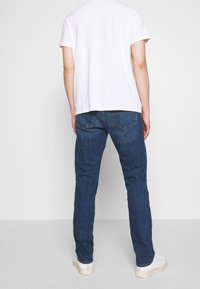 Camiseta blanca ligera, jeans de pierna recta azul oscuro y zapatillas blancas. El denim tiene un lavado ligeramente desgastado y bolsillos traseros visibles.
