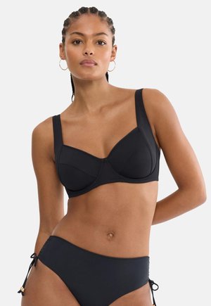 Nő fonott hajjal, fekete merevítős melltartóval és oldalán megkötős bikinialsóval, előre nézve, egyszerű háttér előtt.